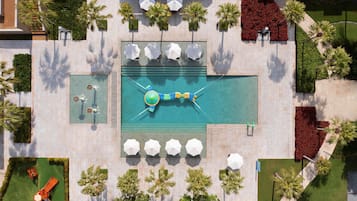An aerial view of a resort with a central swimming pool, multiple lounge areas with umbrellas, and surrounding landscaped gardens.