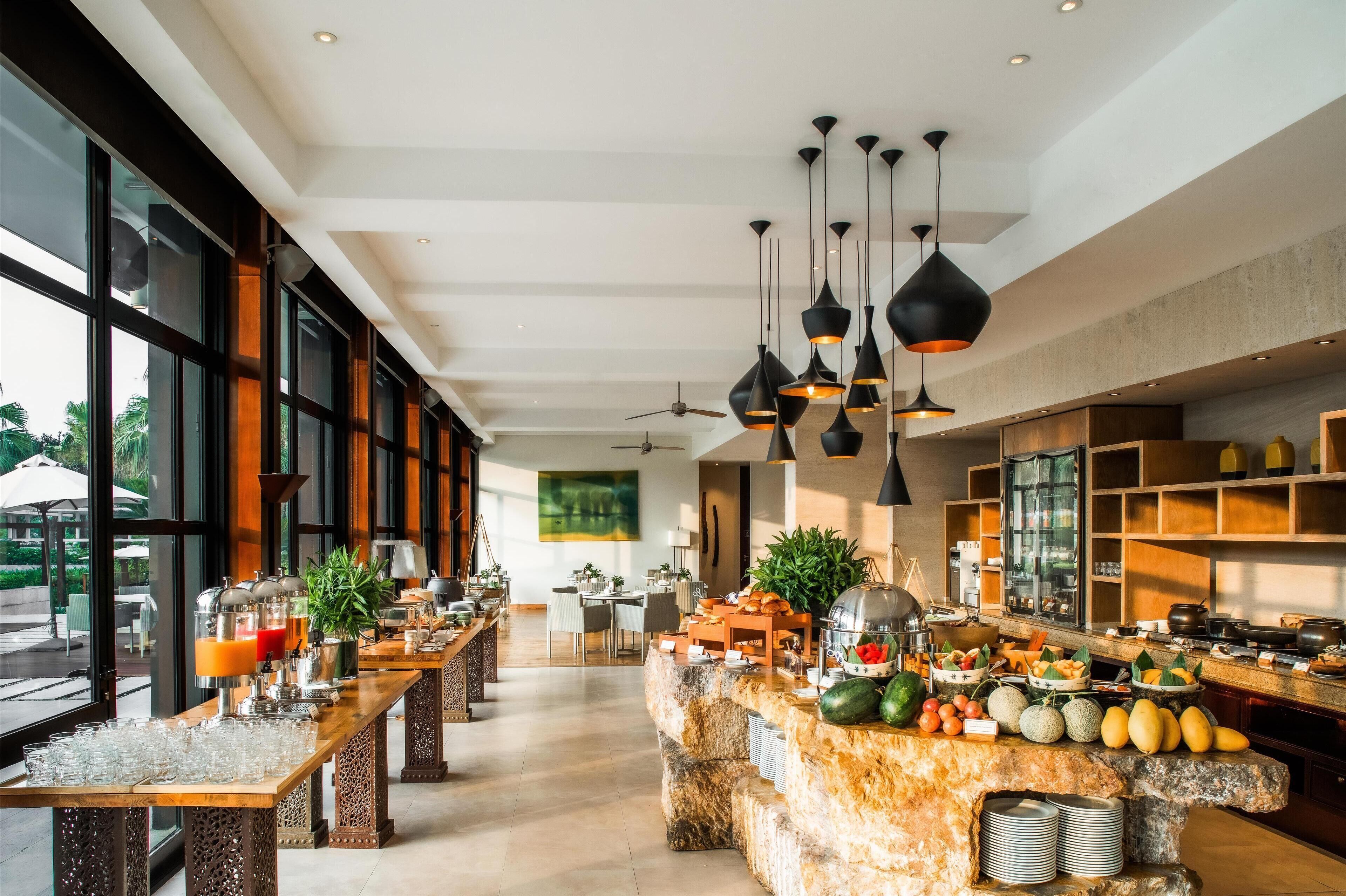 Un amplio vestíbulo de hotel con un mostrador de recepción alargado, ventanas grandes y una cocina moderna con diversos alimentos a la vista.