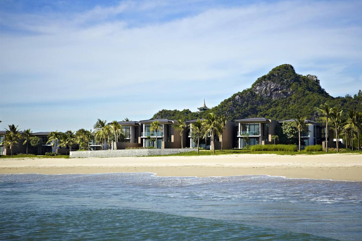 A beachfront hotel with modern architecture, a sandy beach, and a mountain in the background.