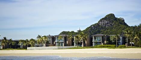 A beachfront hotel with modern architecture, a sandy beach, and a mountain in the background.