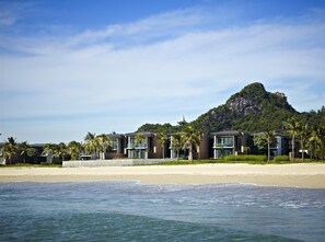 Un hotel frente a la playa con arquitectura moderna, una playa de arena y una montaña al fondo.