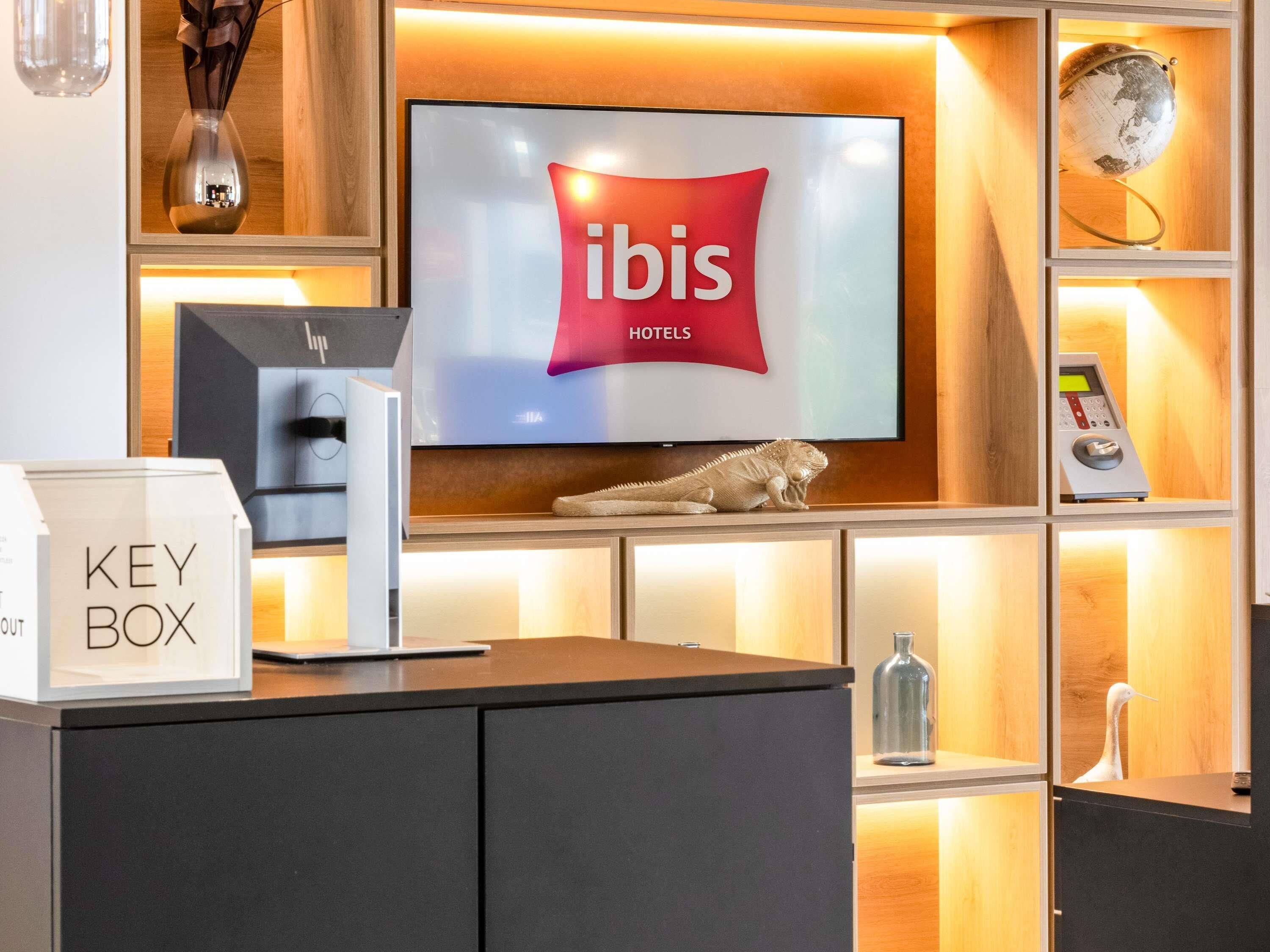 A hotel reception desk with a television displaying the ibis hotels logo, a key box, a digital check-in kiosk, and a decorative iguana.