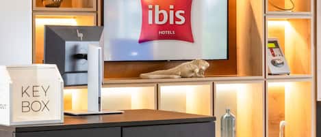A hotel reception desk with a television displaying the ibis hotels logo, a key box, a digital check-in kiosk, and a decorative iguana.