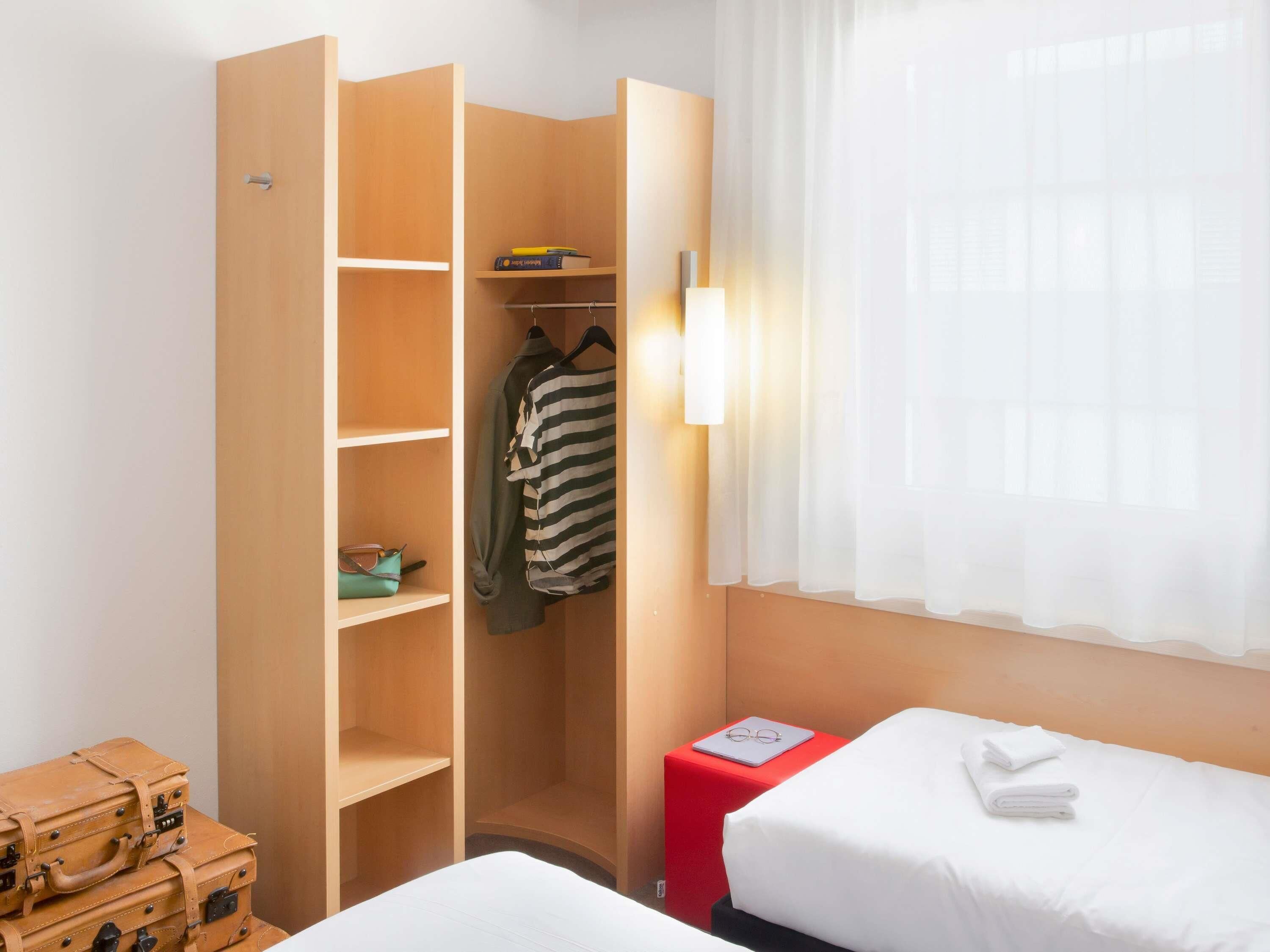 A hotel room with a wardrobe, a bed, a red stool, and a window with sheer curtains.
