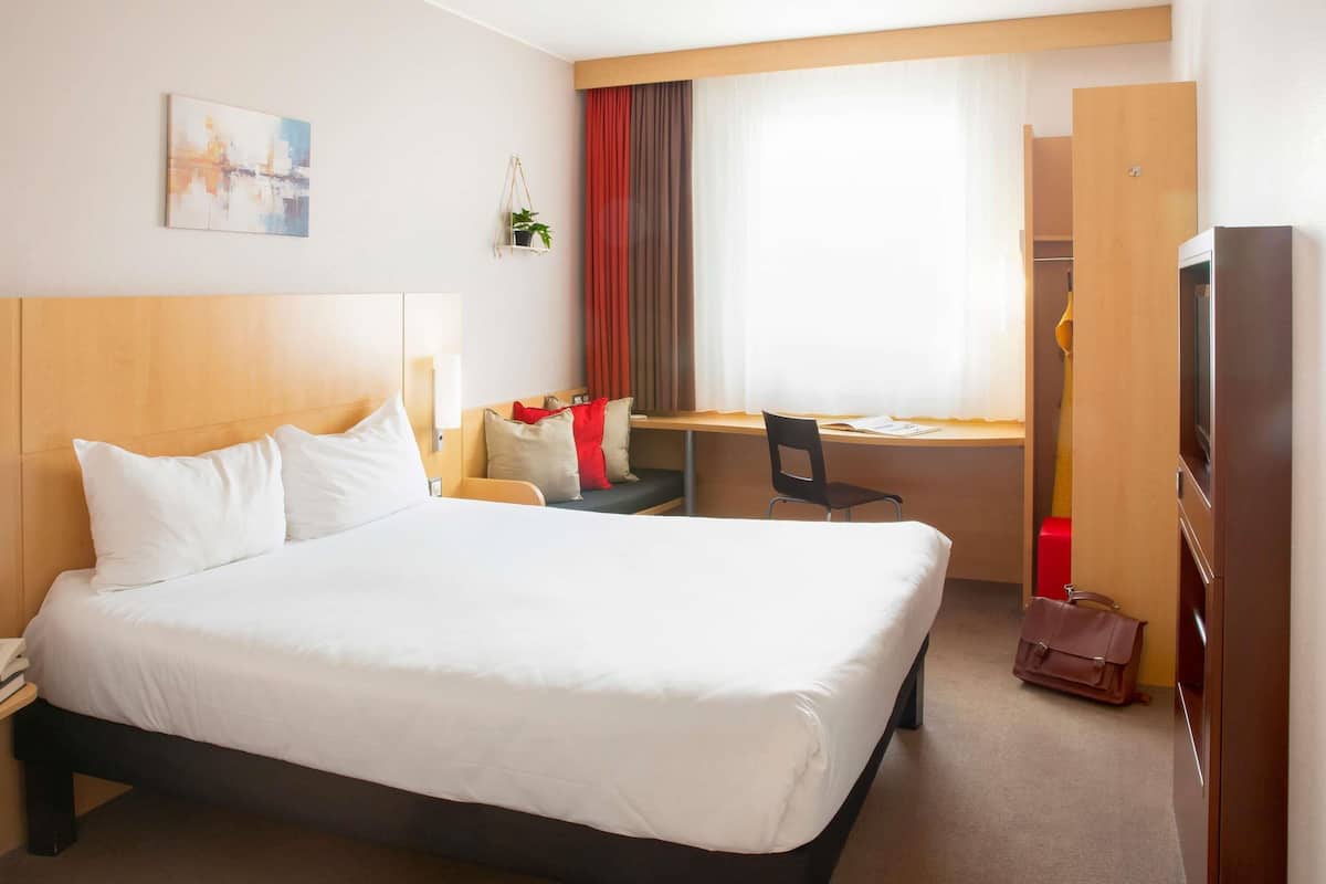 A hotel room with a large bed, a desk with a chair, a TV, and a wardrobe.