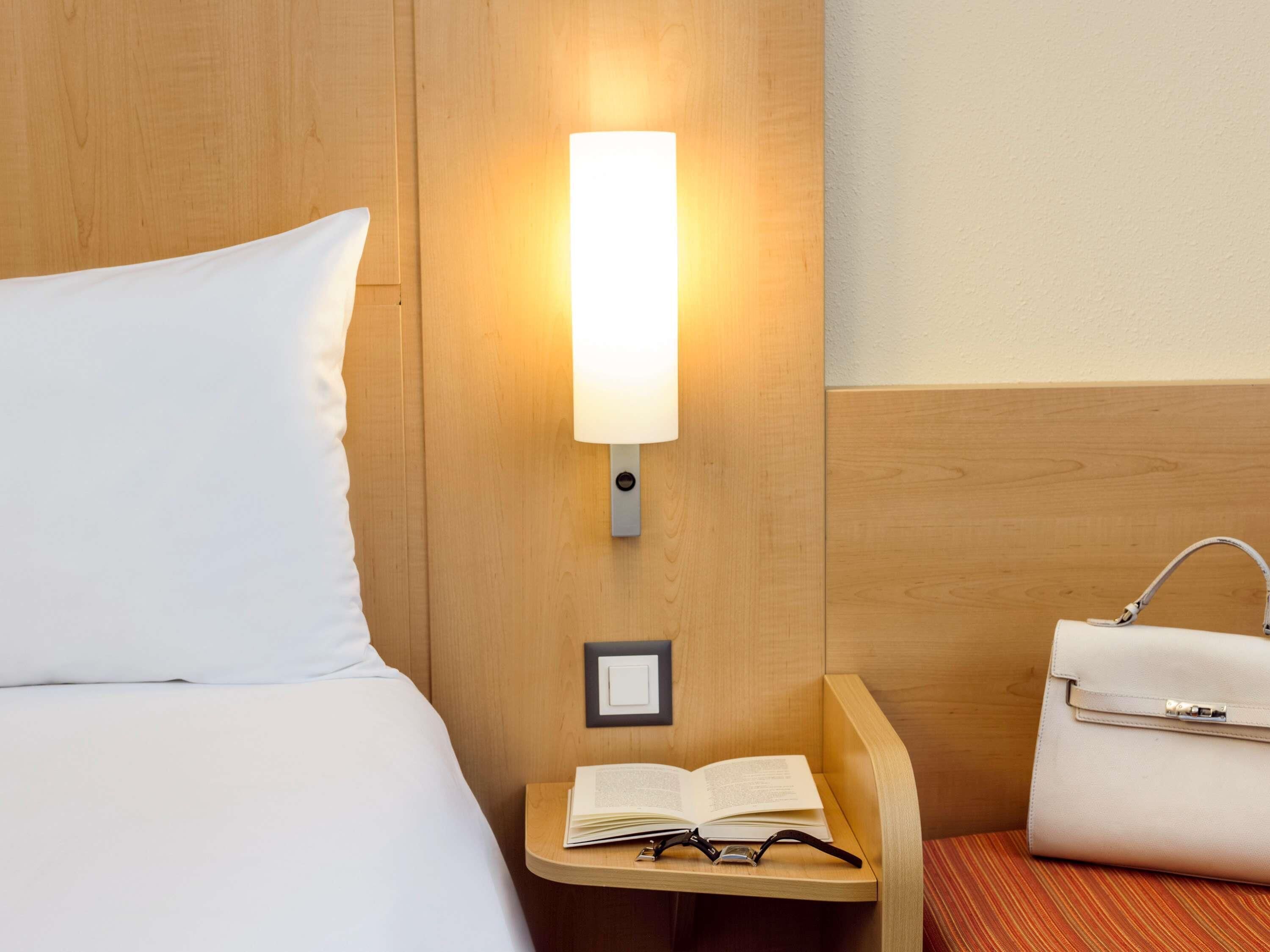 Habitación de hotel con cama, mesita de noche con lámpara, un marco de fotos pequeño, un libro abierto y un bolso.