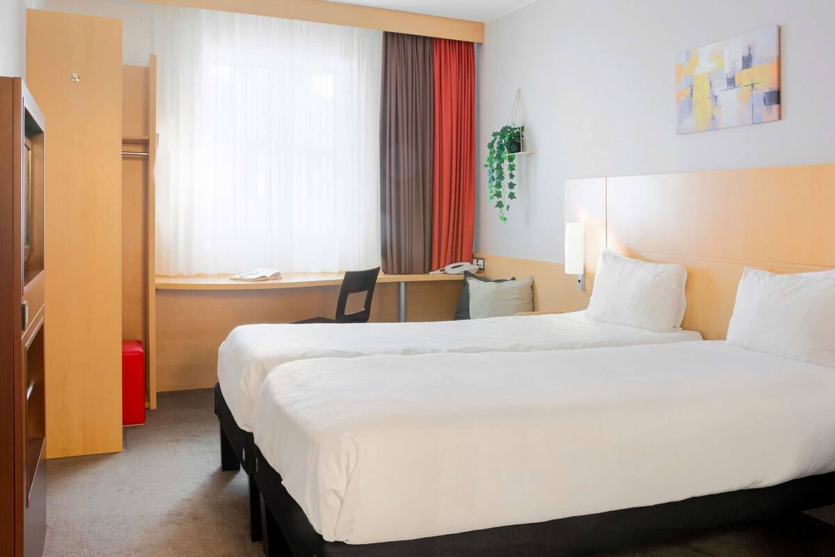 A hotel room with two beds, a desk, a chair, a window with curtains, and a wall-mounted plant.