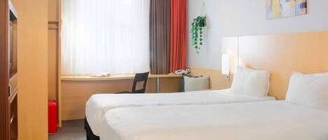 A hotel room with two beds, a desk, a chair, a window with curtains, and a wall-mounted plant.