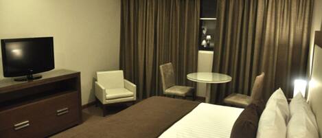 Habitación de hotel con una cama, un televisor sobre una cómoda de madera, una silla, una mesita y una ventana con cortinas.