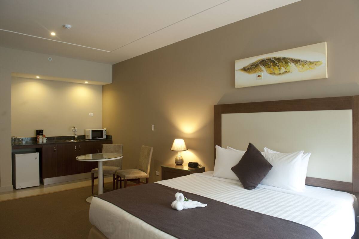 A hotel room with a bed, a desk, a chair, a microwave, a lamp, and a painting on the wall.