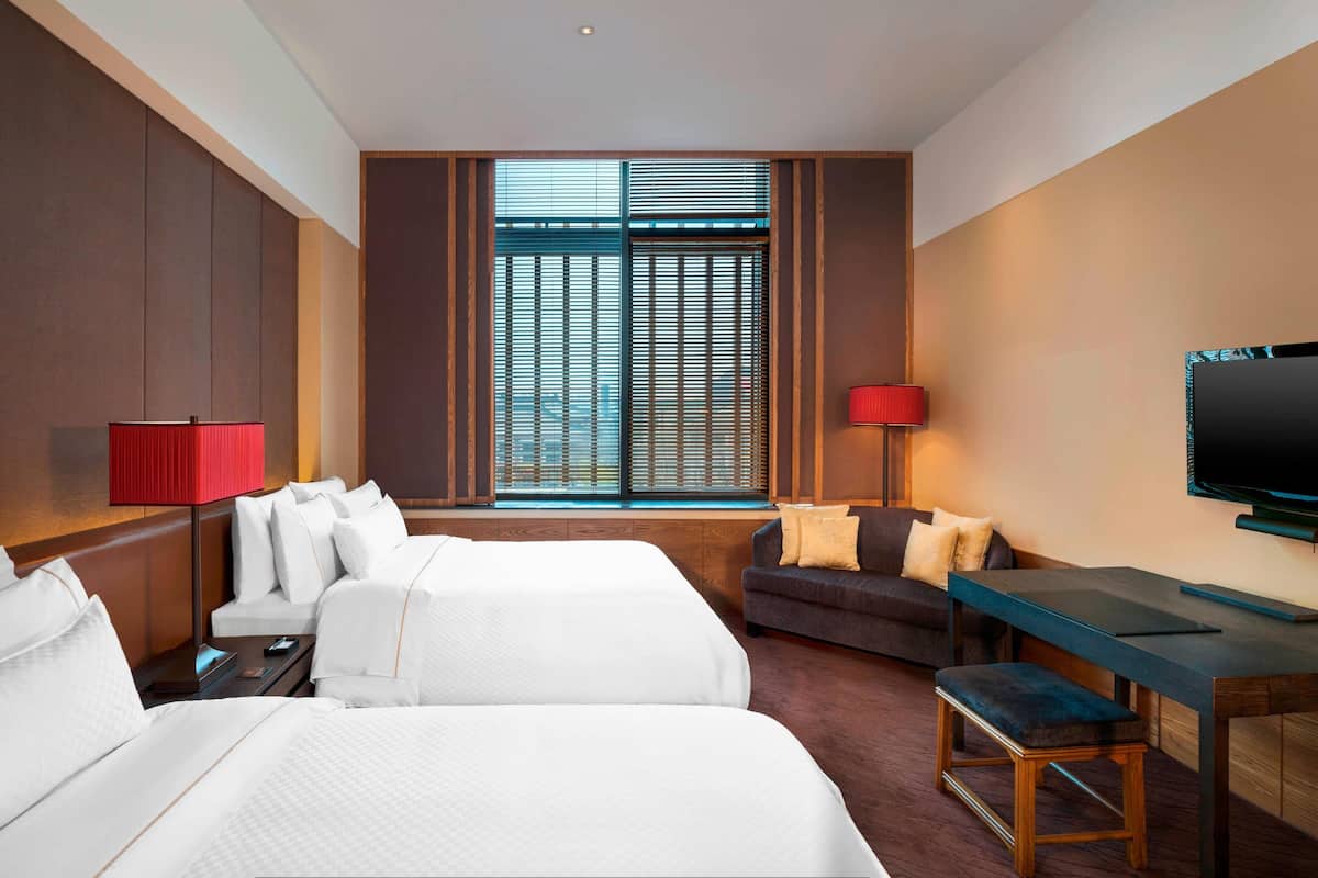 A hotel room with two beds, a sofa, a desk, a TV, and a window with blinds.