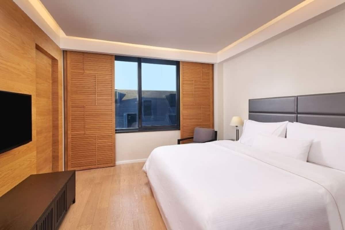 A modern hotel room with a large bed, a flat-screen TV, wooden paneling, and a view of the city.