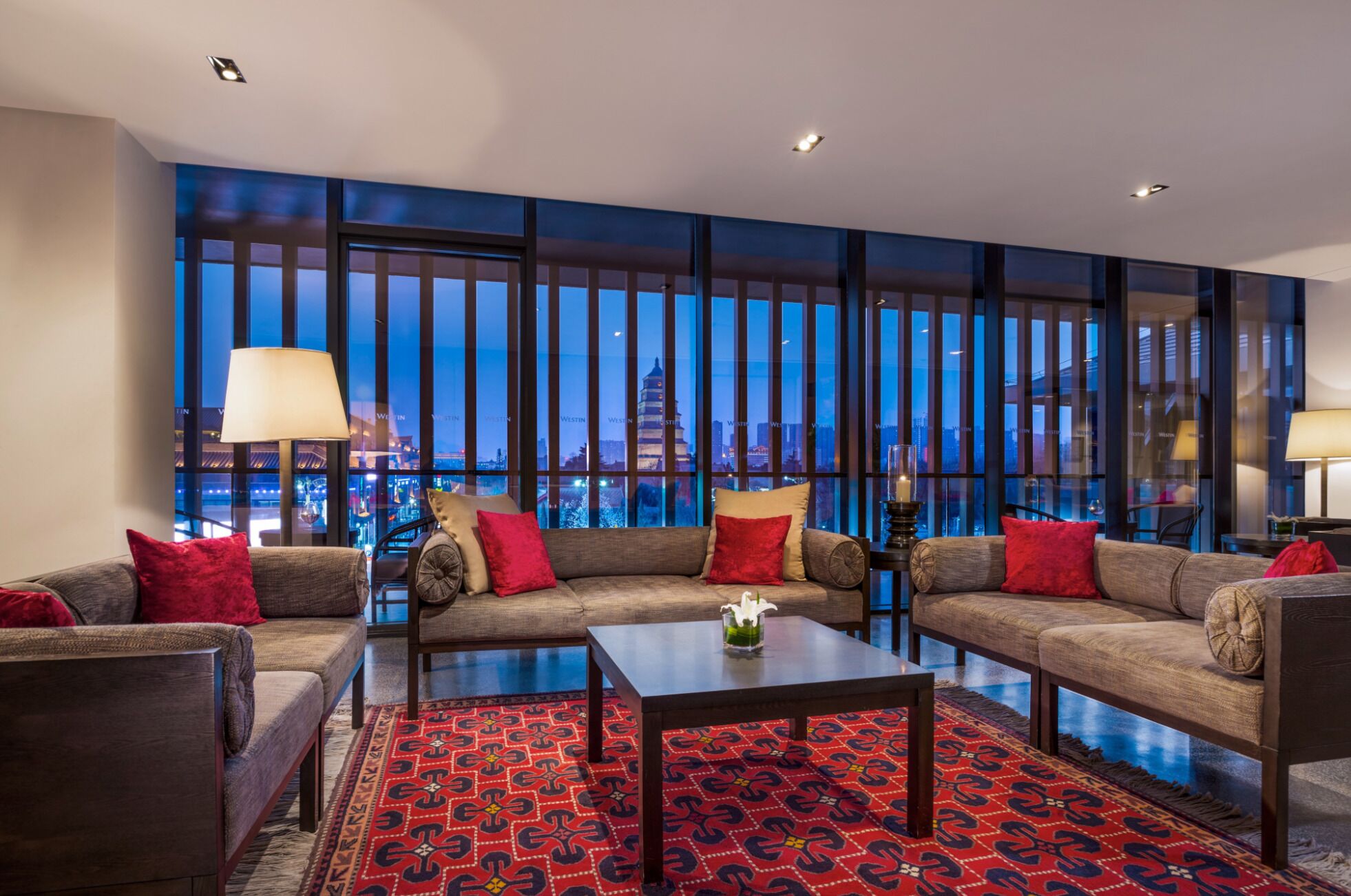 A modern hotel room with a large window, a sofa set, a coffee table, and a patterned rug.