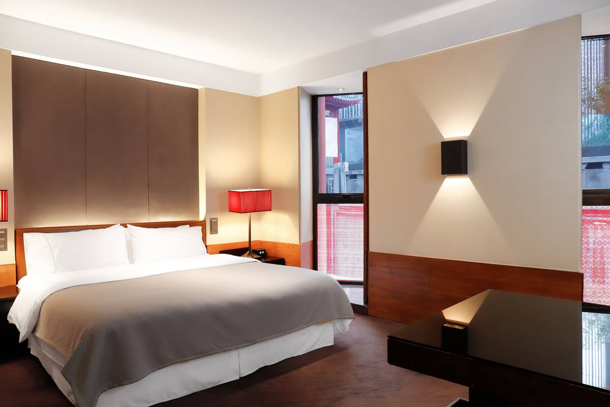 A hotel room with a large bed, a bedside table with a lamp, a wall-mounted TV, and a window with a view of buildings.