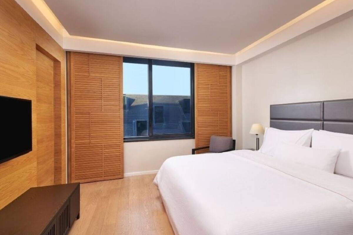 A modern hotel room with a large bed, a flat-screen TV, wooden paneling, and a view of the city.