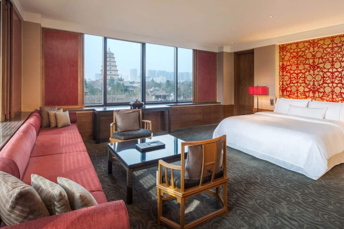 A hotel room with a large bed, a red sofa, a wooden chair, a small table, and a view of a cityscape through large windows.