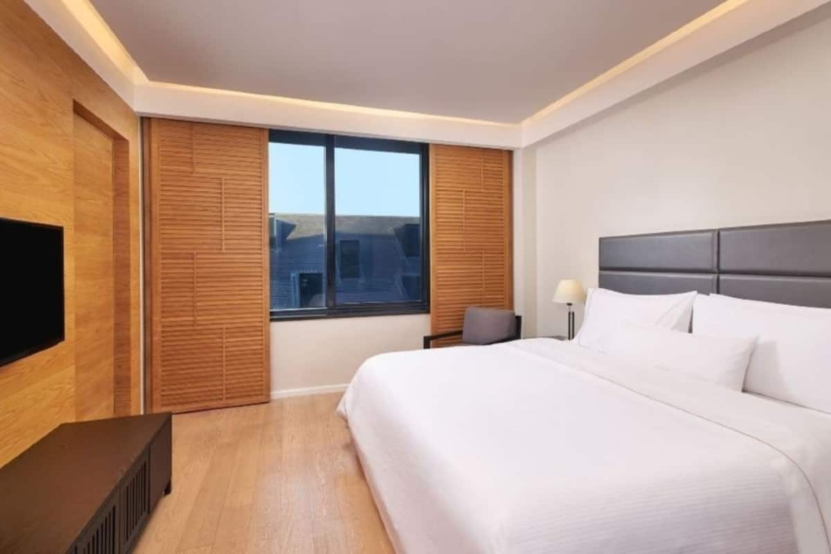 A modern hotel room with a large bed, wooden paneling, a television, and a view of the cityscape.