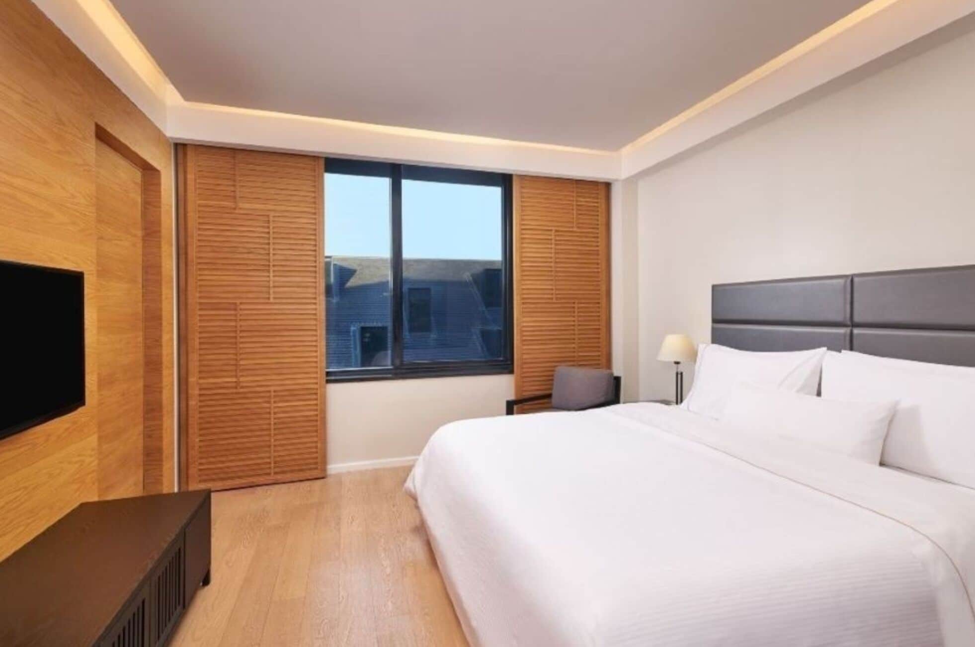 A hotel room with a large bed, a television, a window with blinds, and a nightstand with a lamp.