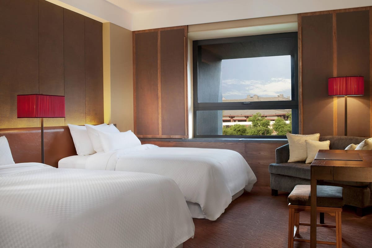 A hotel room with two beds, a desk, and a chair. There is a large window providing a view of buildings and greenery.