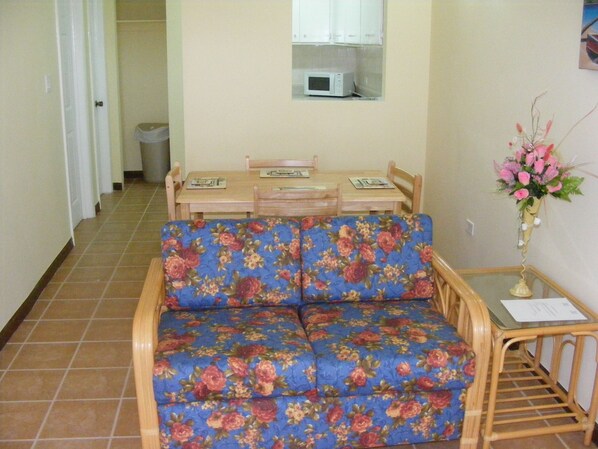 A living room with a floral sofa, a dining table with chairs, a microwave above the table, and a vase with flowers on a side table.