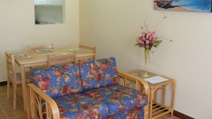 Una habitación con una mesa de comedor de madera, dos sillas, un sofá con funda floral, una pequeña mesita con un jarrón y un cuadro de una playa, además de un espejo en la pared.
