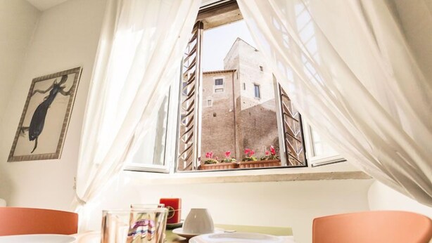 A dining area with a table set for two, a window with curtains, and a view of a building outside. - Maison Colosseo (Rome)