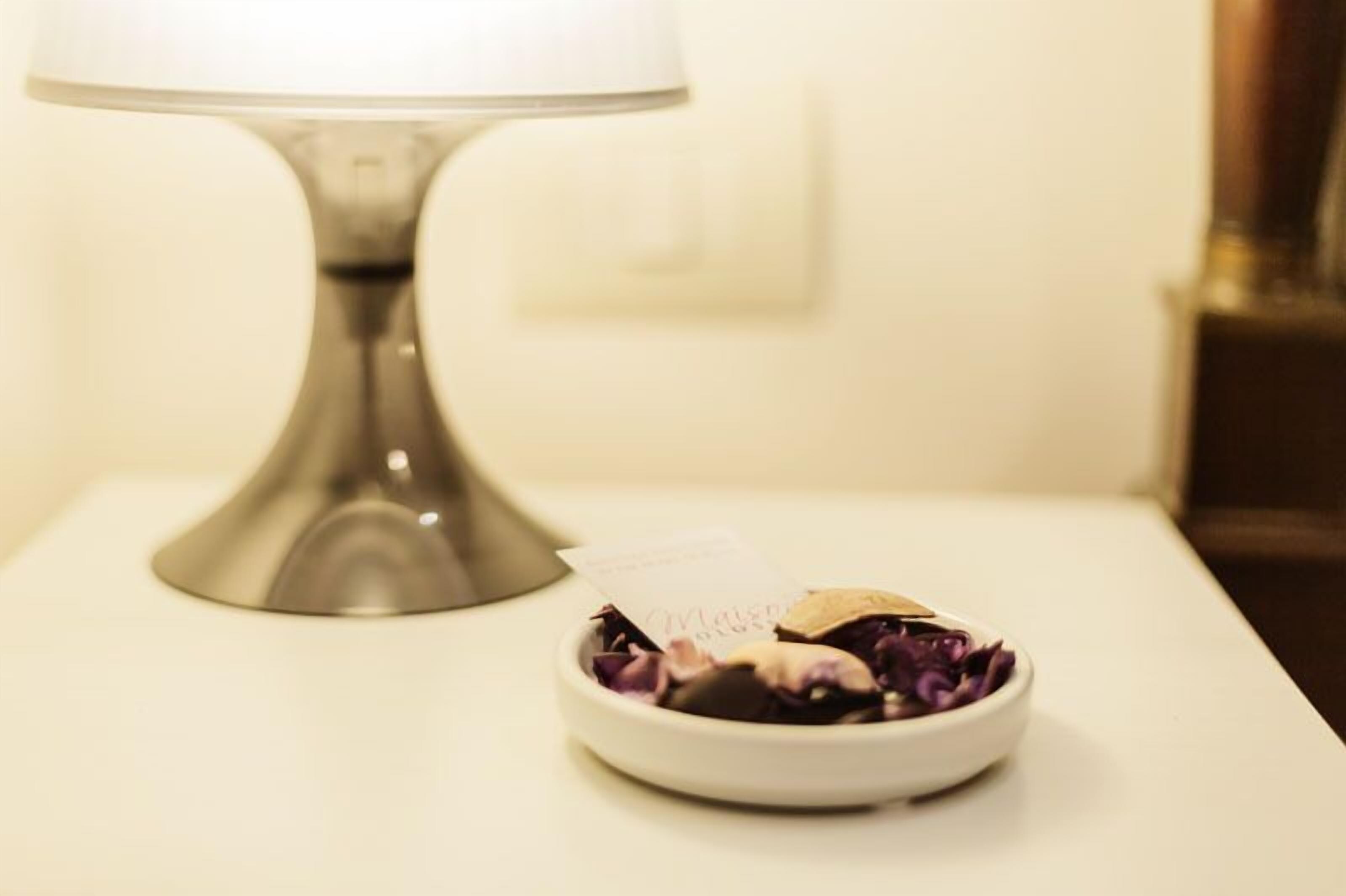 A white bedside table with a metal lamp and a bowl containing purple flowers.