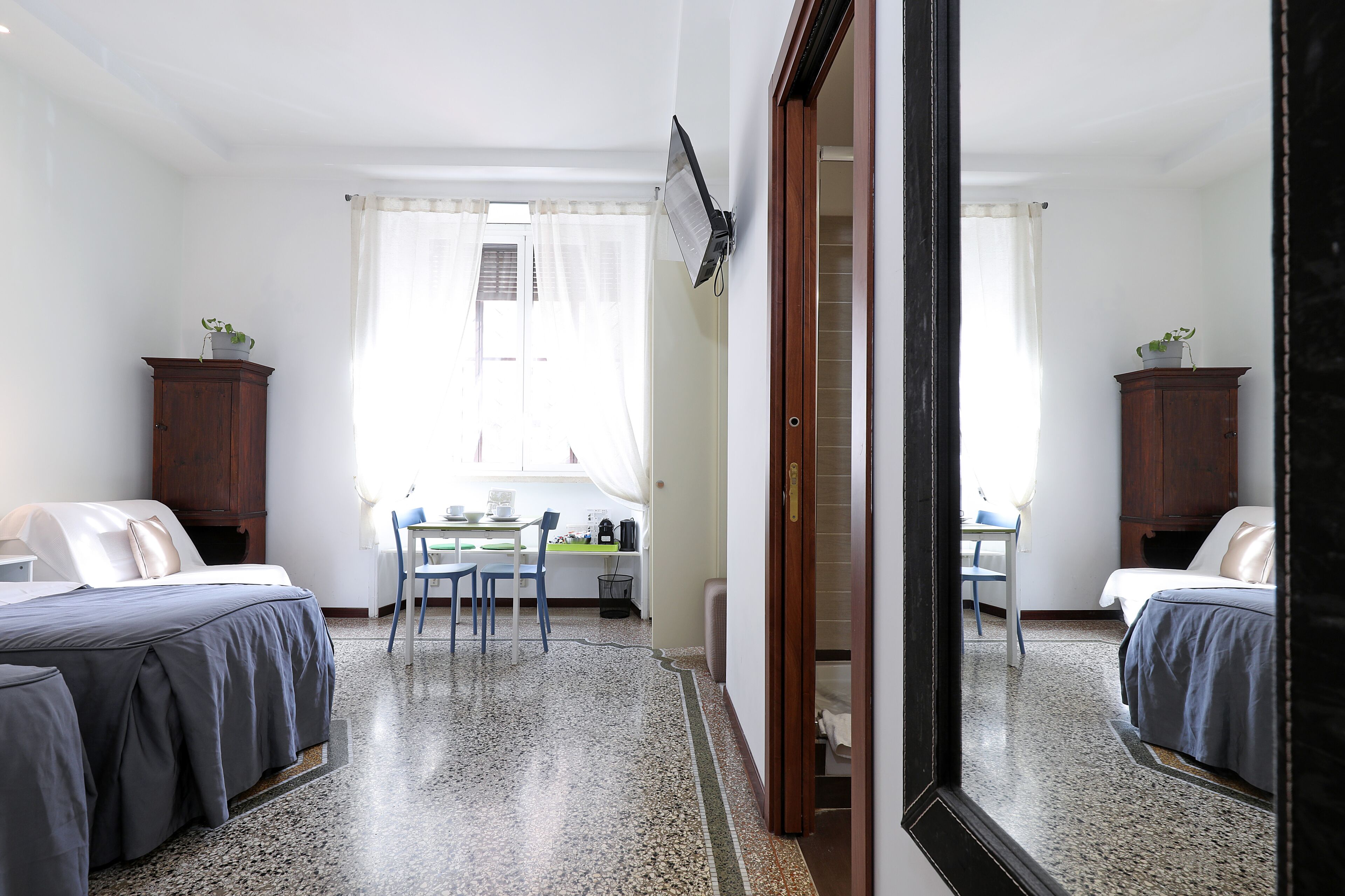 A bedroom with two beds, a desk, chairs, a mirror, and a window with curtains.