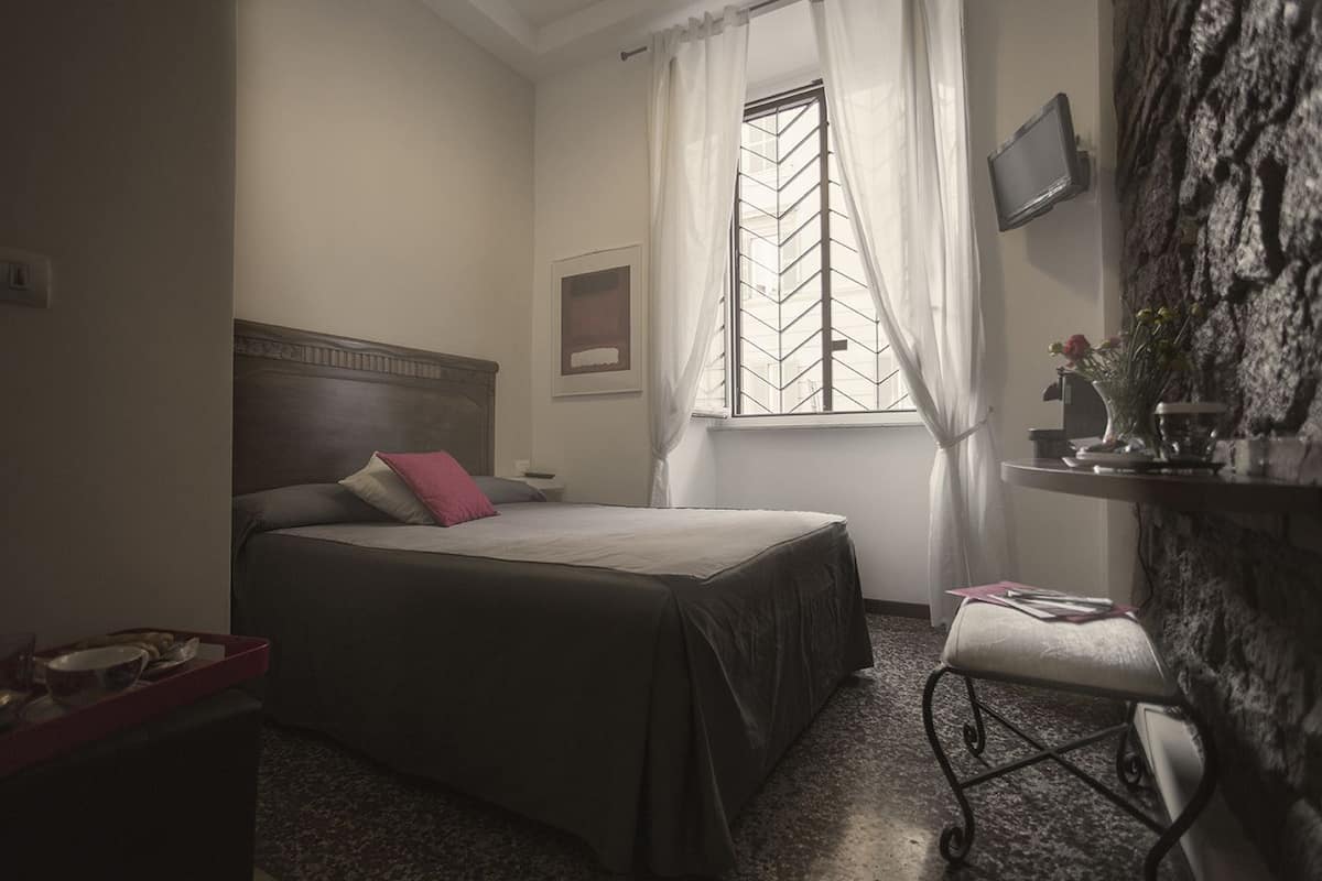 A bedroom with a bed, a nightstand, a window with curtains, a wall-mounted TV, and a small table with a vase of flowers.