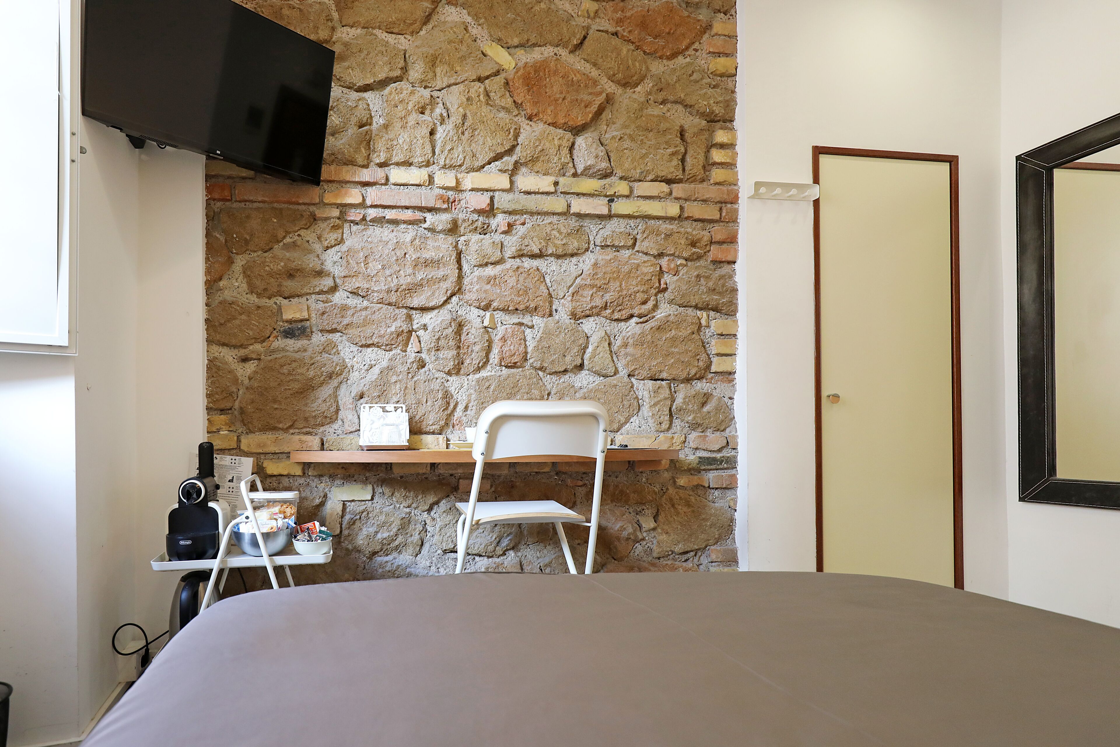 Une pièce avec un mur en pierre, un téléviseur à écran plat fixé au-dessus d’un bureau, une chaise et un lit.