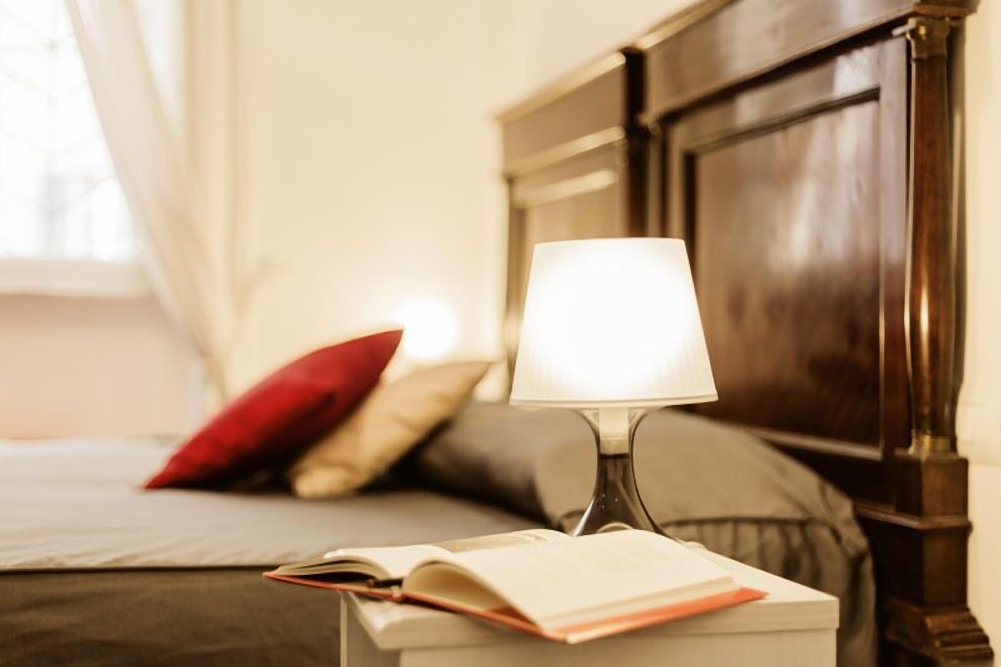 A neatly made bed with a red pillow, a bedside lamp, and an open book on a nightstand.