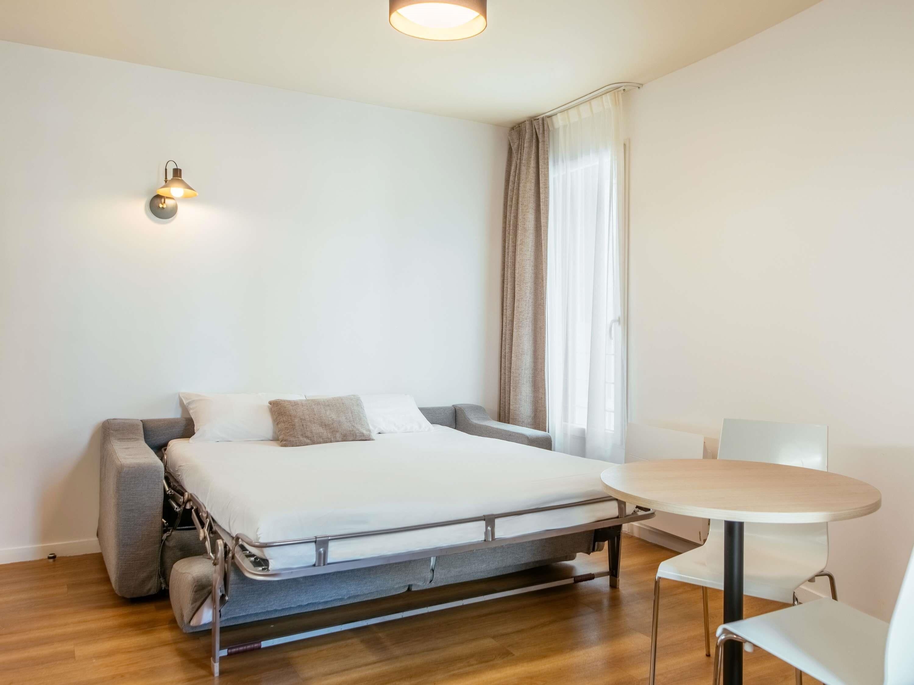 Een kleine kamer met een bed, een tafeltje en een lampje aan de muur.