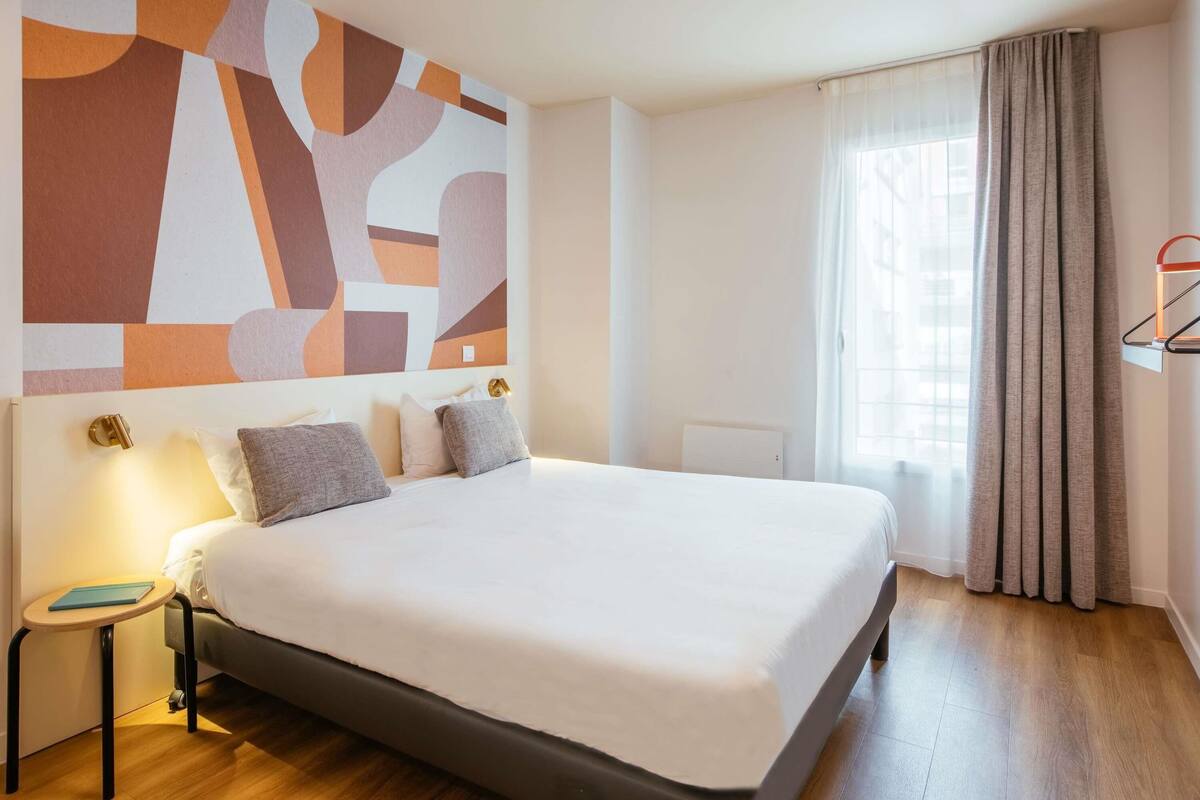 Une chambre d’hôtel avec un grand lit, des tables de chevet, une œuvre d’art murale, une fenêtre avec des rideaux et un radiateur.