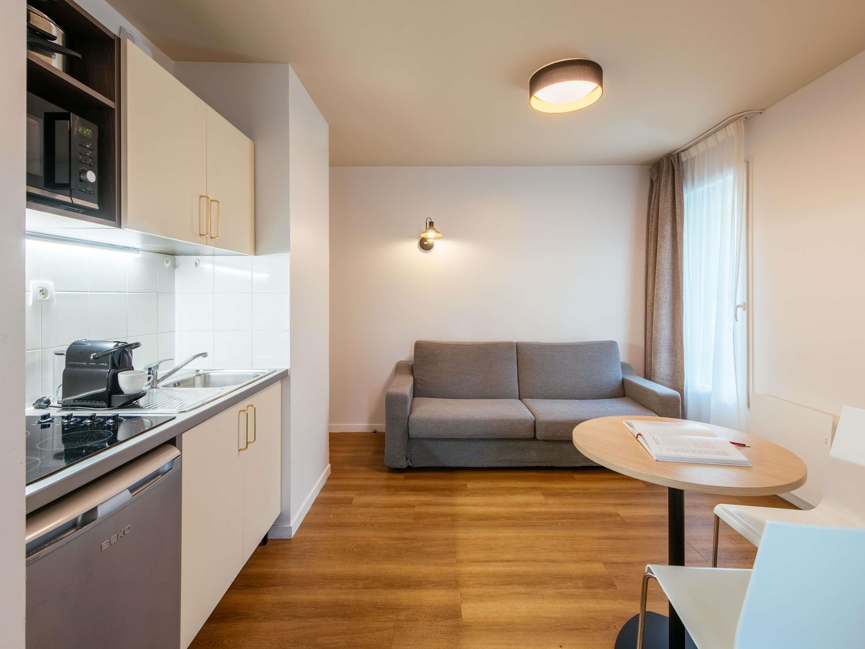 Una cocina compacta con microondas, tostadora y lavamanos. En la sala de estar, un sofá gris y una pequeña mesa redonda con un libro.