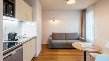 Una cocina compacta con microondas, tostadora y lavamanos. En la sala de estar, un sofá gris y una pequeña mesa redonda con un libro.