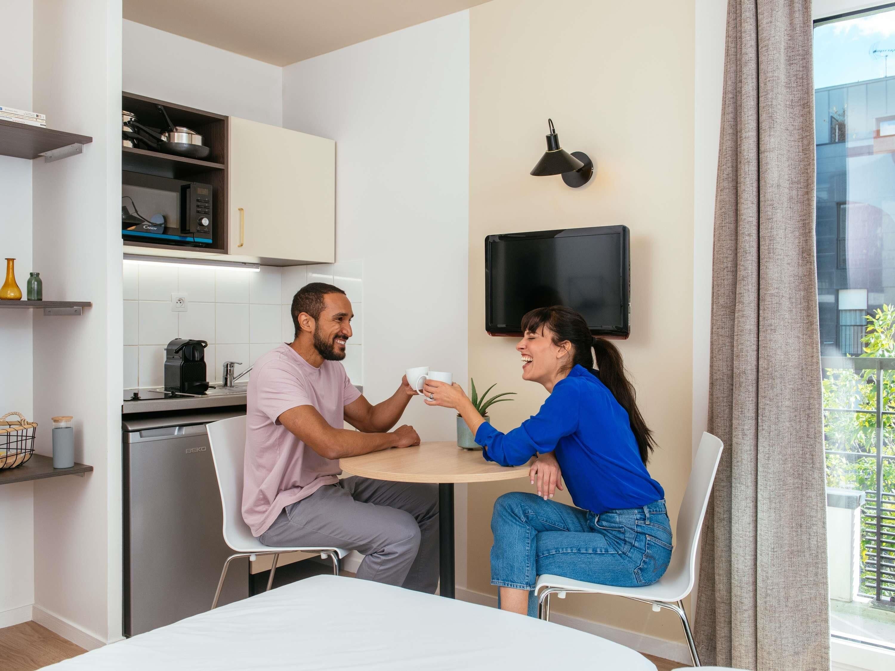 A modern kitchen with a refrigerator, microwave, and coffee maker, a dining area with a table and chairs, and a TV mounted on the wall.