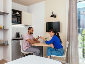 A modern kitchen with a refrigerator, microwave, and coffee maker, a dining area with a table and chairs, and a TV mounted on the wall.