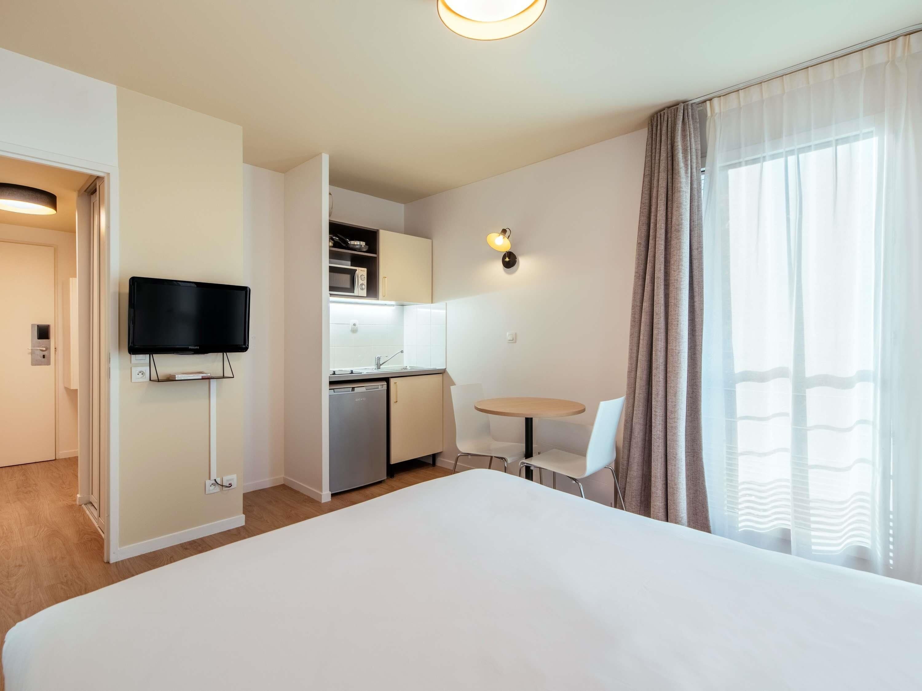 Ein Hotelzimmer mit einem Bett, einem an der Wand befestigten Fernseher, einer kleinen Küchenzeile inklusive Mikrowelle, Spüle und Geschirrspüler. Ein runder Tisch mit zwei Stühlen und ein Fenster mit Vorhängen sind ebenfalls vorhanden.