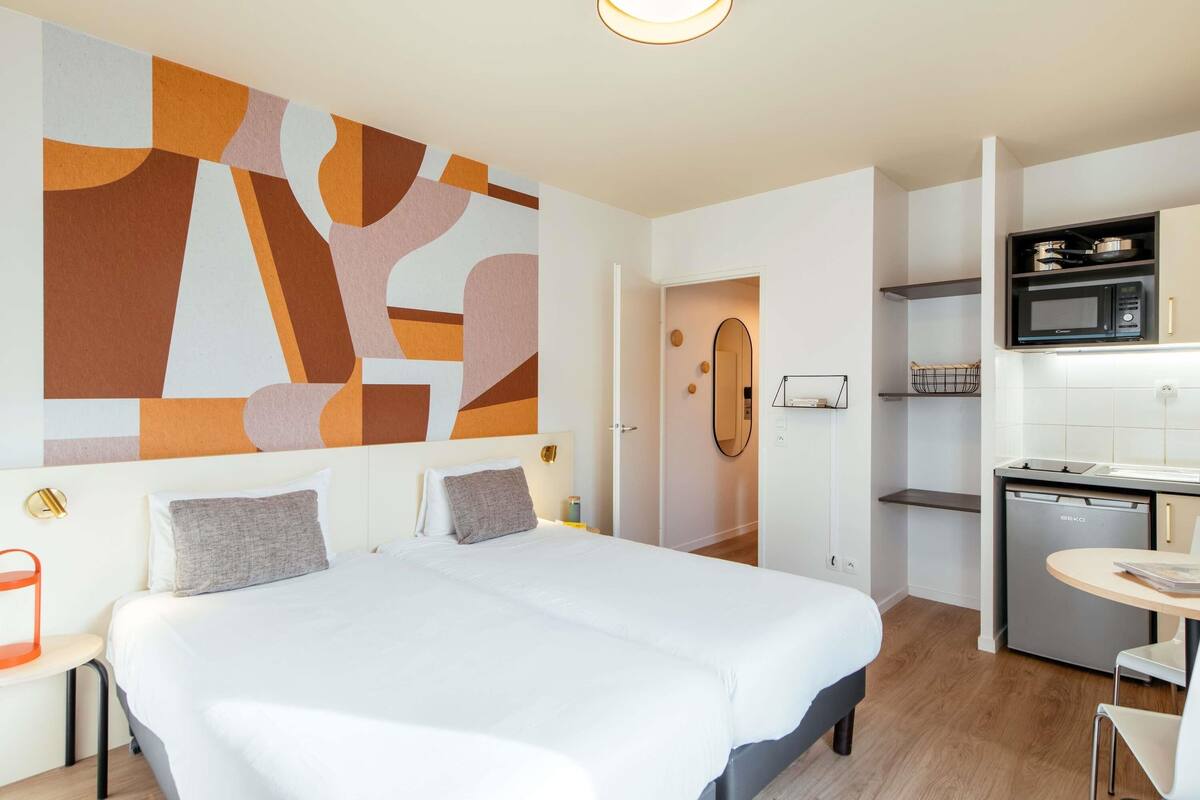 Une chambre d’hôtel moderne avec un grand lit, une kitchenette et une salle de bain visible à travers une porte ouverte.