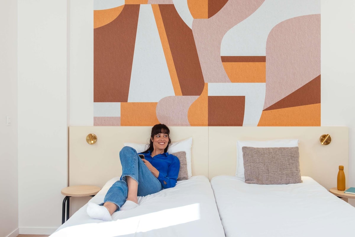 Une personne allongée sur un lit, un téléphone intelligent à portée de main, une grande peinture abstraite accrochée au mur et une petite table ronde sur laquelle se trouve une bouteille.