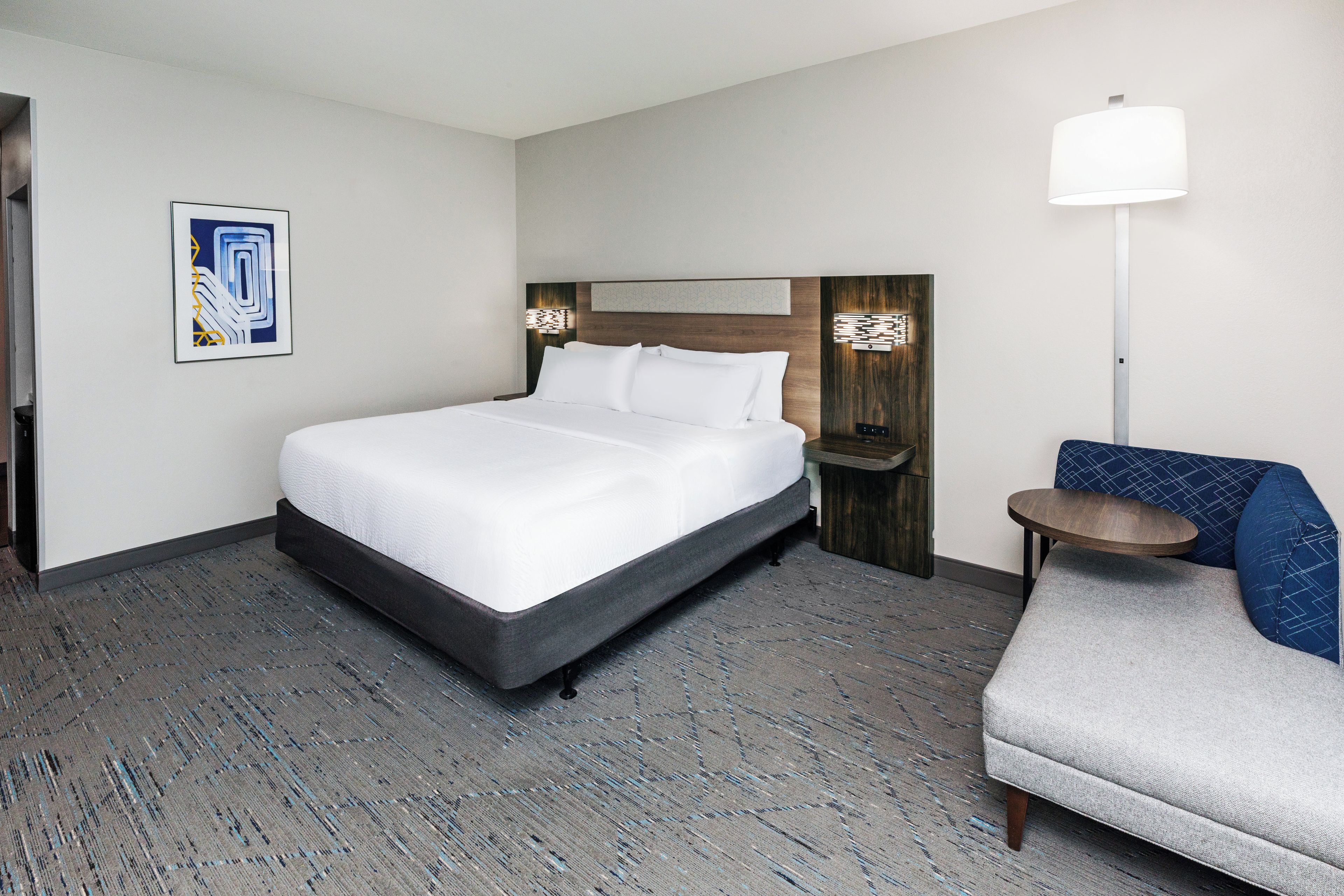 A hotel room with a bed, a chair, a nightstand, a lamp, and a wall-mounted light fixture.