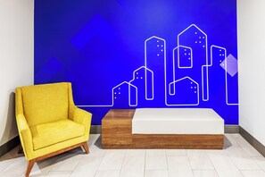 Une pièce avec un fauteuil jaune, un banc en bois et un mur bleu orné d’un motif géométrique représentant un paysage urbain.