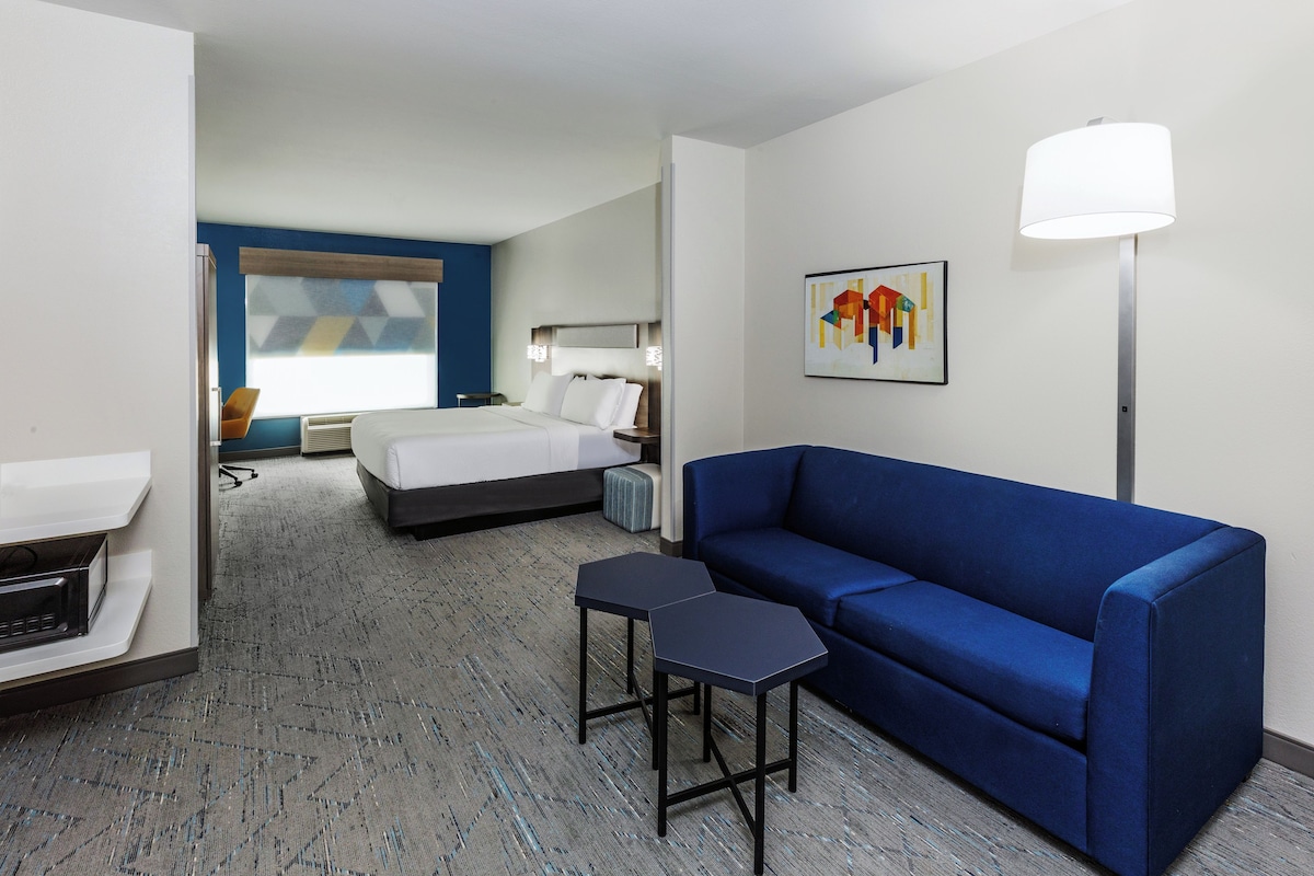 A hotel room with a bed, a blue sofa, a small table, a desk, a chair, a lamp, and a painting on the wall.