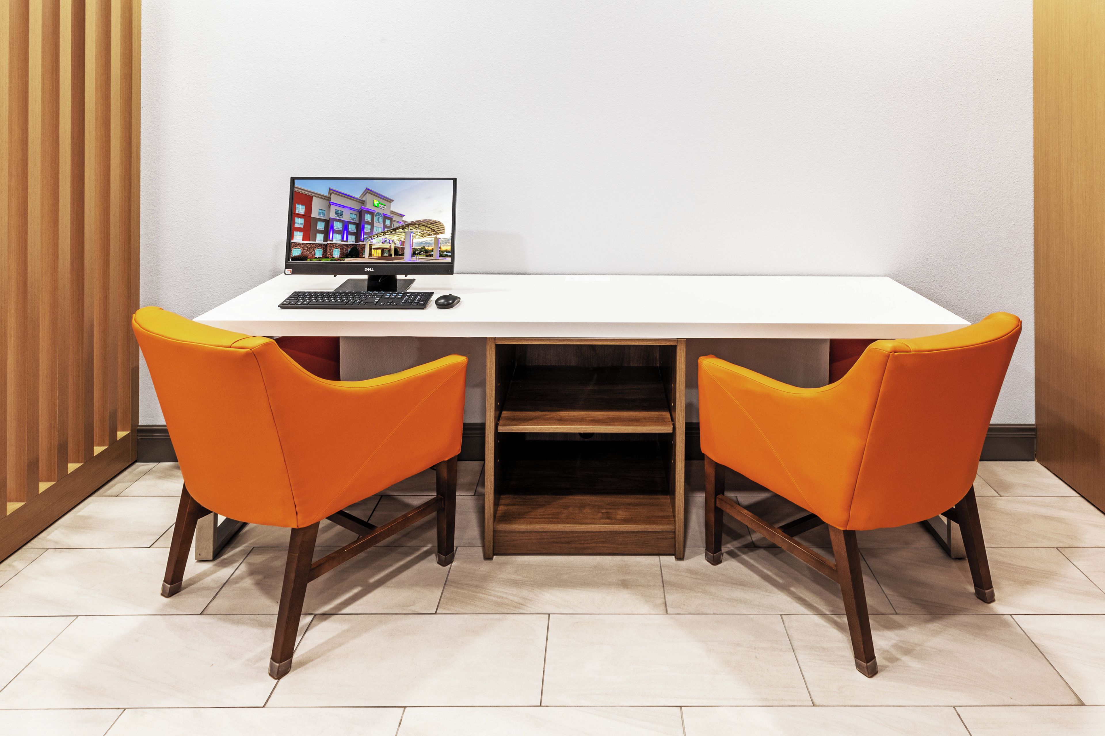 A hotel room with a white desk, two orange chairs, a Dell monitor, and a keyboard.