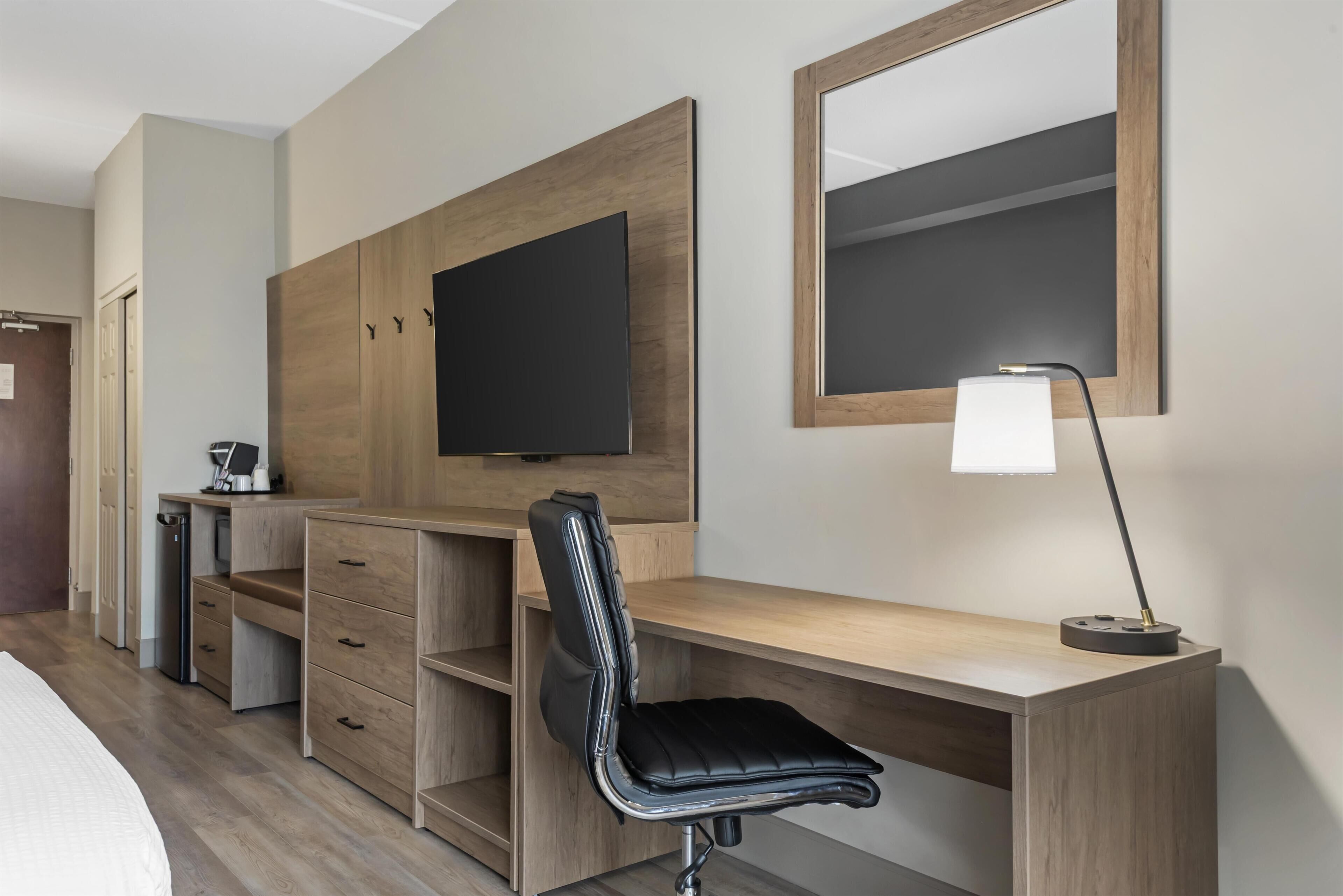 Quarto de hotel com escrivaninha de madeira, cadeira de escritório preta, televisão fixada na parede e abajur.