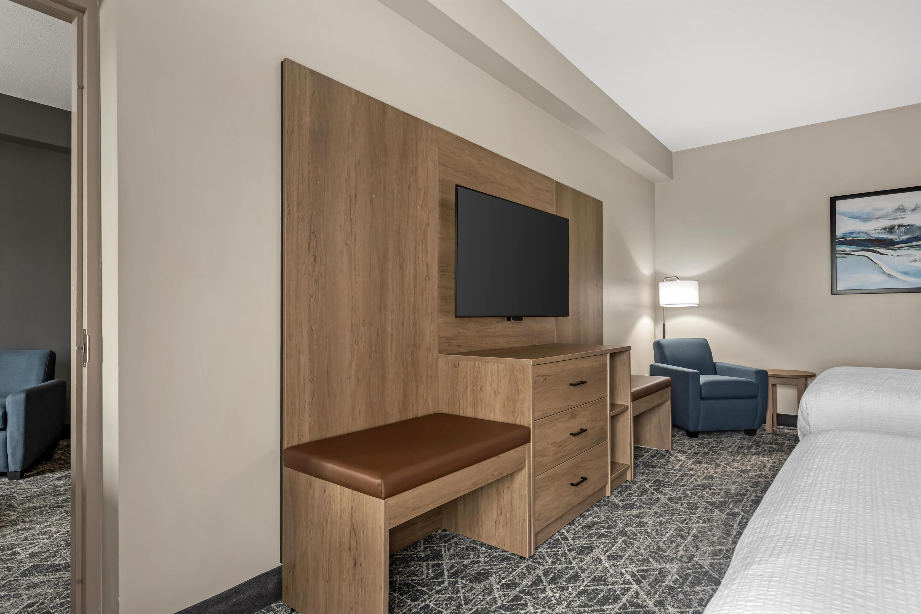 Habitación de hotel con una cama, un escritorio con cajones, una silla, un televisor montado en la pared y una mesita de noche con lámpara.