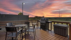An outdoor deck with a grill, chairs, and a table, overlooking a sunset.