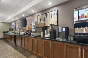 Zona de desayuno de hotel con una barra larga, máquinas de café y un refrigerador.