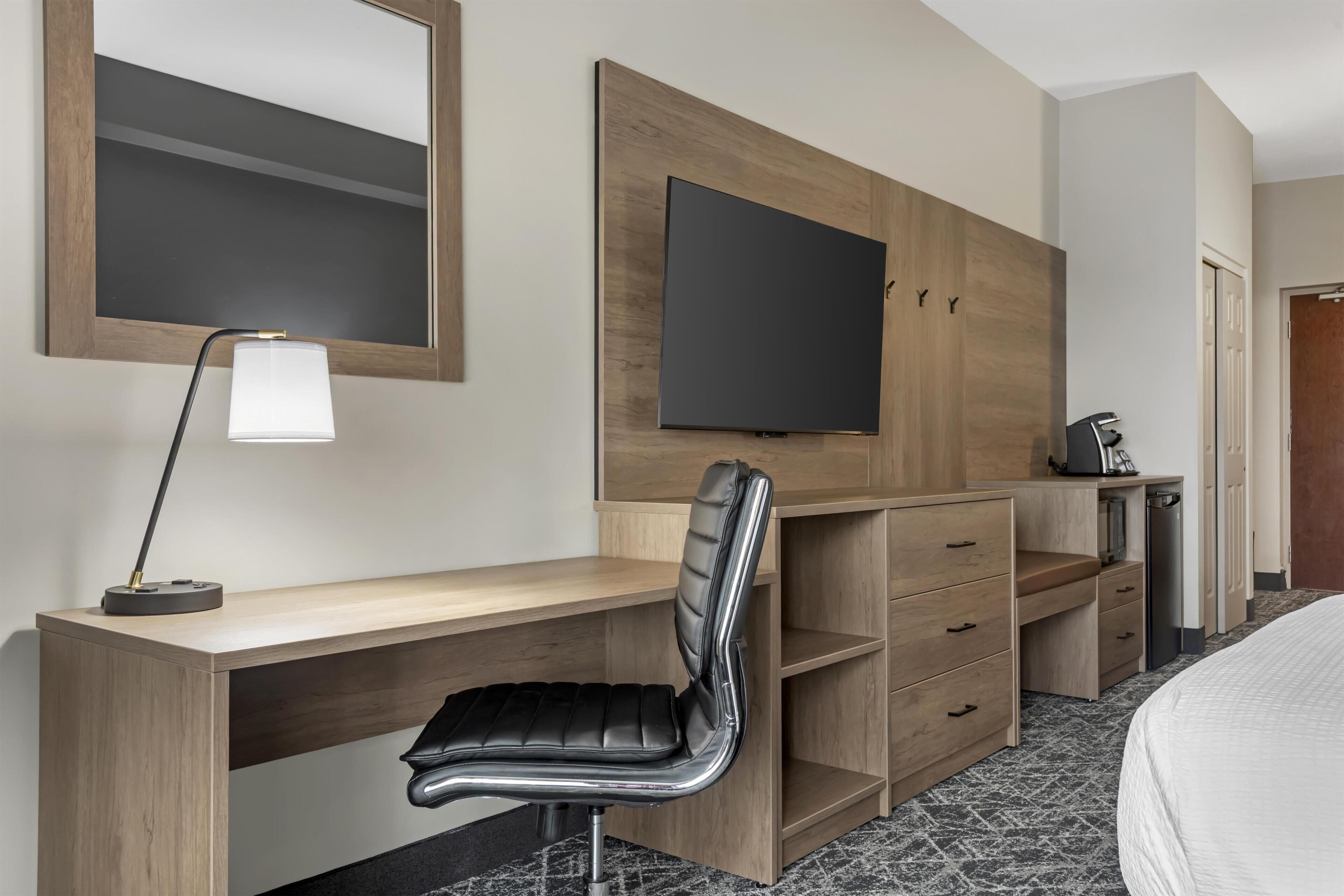 A hotel room with a wooden desk, a chair, a television mounted on the wall, and a bed with a white bedspread.