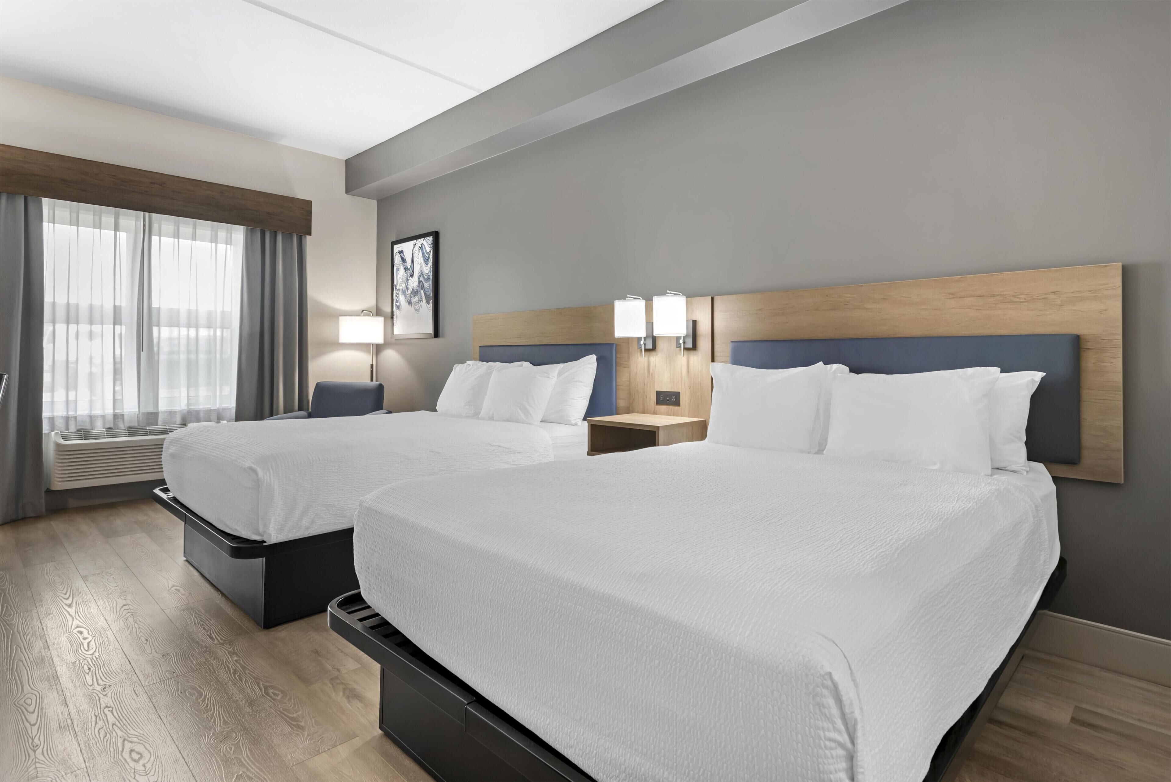 Een hotelkamer met twee bedden, een groot raam met gordijnen, een kunstwerk aan de muur en een nachtkastje met een lamp.